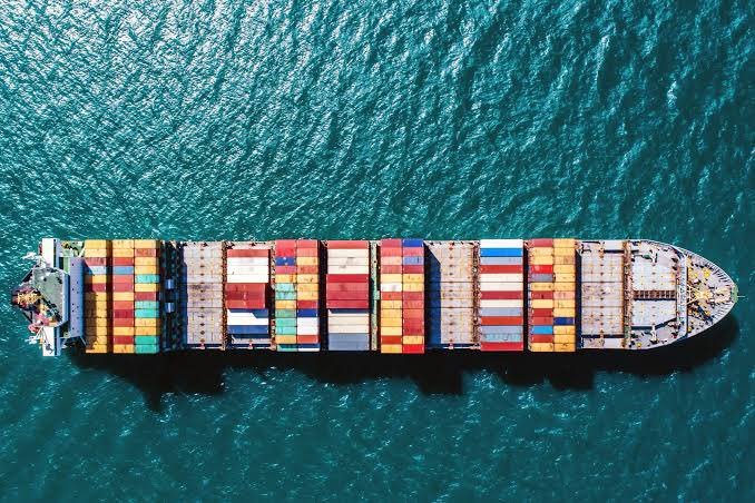 An aerial top-down view of a large cargo ship loaded with colorful shipping containers floating in turquoise water.