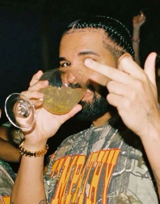 A man with braided hair and a beard drinking from a wine glass with a small dark object inside, while making hand gestures towards the camera.