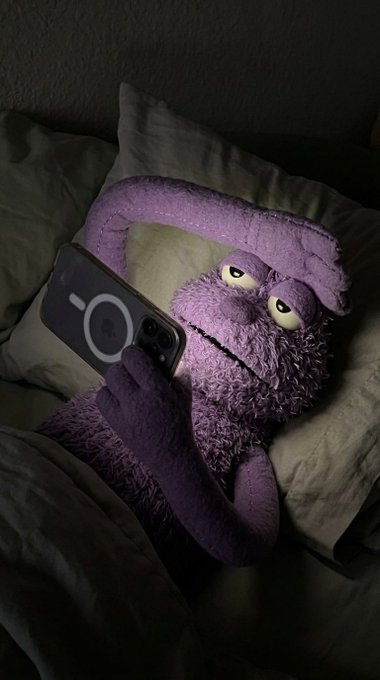 A purple puppet with tired eyes is in bed, holding an iPhone with a MagSafe charger visible. The puppet's posture and the phone in hand visually represent the act of constantly checking the $ANSEM stock chart.