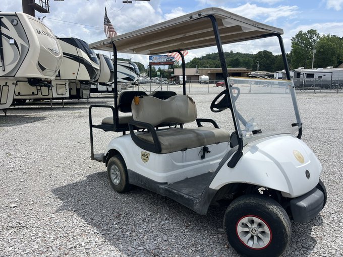 A white Yamaha golf cart with tan seats is parked on gravel at a dealership, with several large white RVs and an American flag visible in the background.