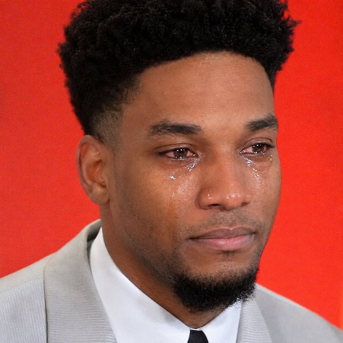 A young man with curly hair and a beard is seen crying, with visible tears rolling down his cheeks, wearing a light gray suit jacket against a solid orange background, humorously used to express the heartbreak of watching a cryptocurrency ($ANSEM) investment plummet in real-time while unable to act on it.