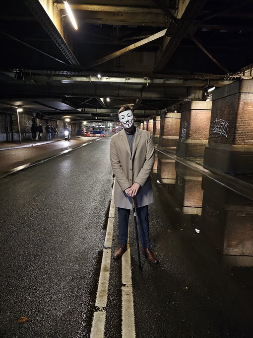 A man wearing a tan coat and a Guy Fawkes (Anonymous) mask stands on a wet road beneath a bridge at night, holding a cane, with brick pillars visible to the side.