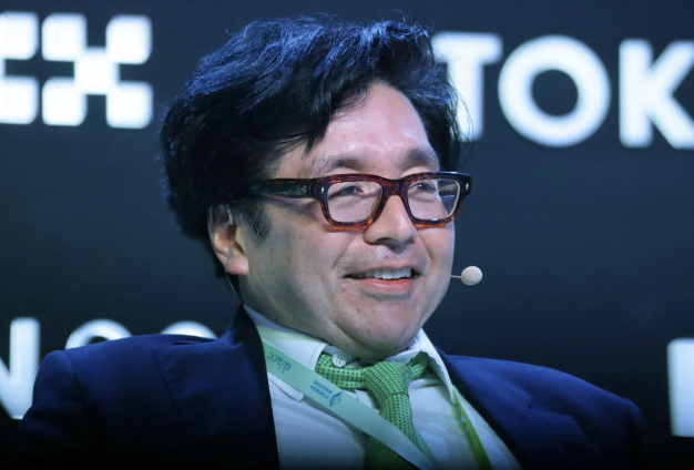 A man in glasses and a green tie smiles while speaking at an event; the partial "TOK" text behind him suggests this is a Tokyo conference appearance. The image accompanies news of Tom Lee calling Robinhood Chain proof that ETH serves as digital money, highlighting Ethereum's native gas and fee denomination.