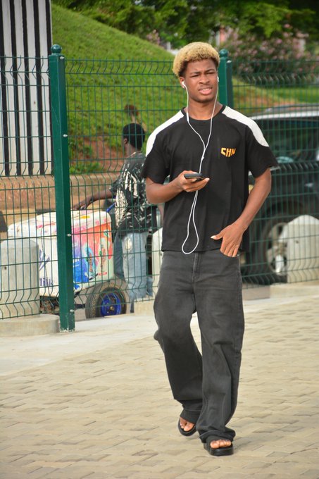 A young man with blonde-tipped hair walks outdoors while listening to white earbuds and looking at his phone.