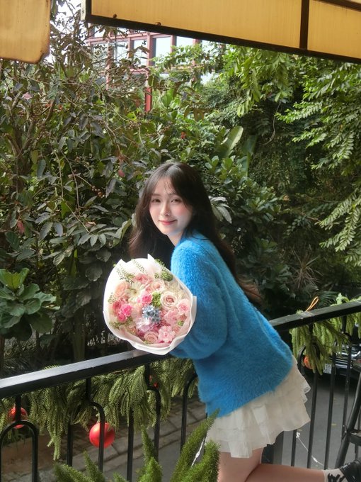 A young woman wearing a blue fuzzy sweater and white skirt, smiling while holding a bouquet of pink and pastel flowers on a balcony surrounded by plants.