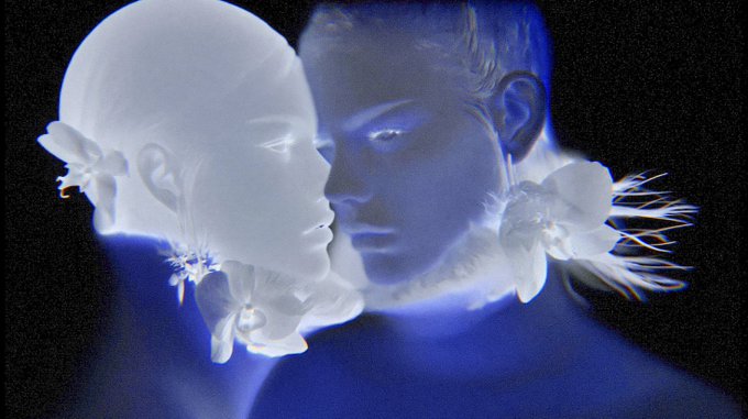 An ethereal, glowing digital artwork featuring two translucent, bluish-white face profiles facing each other, adorned with flower shapes, against a dark background.