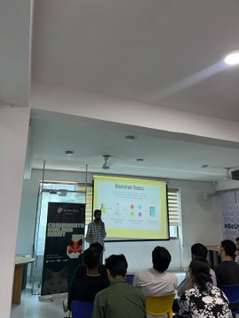 A photograph of a classroom or conference room where a presenter stands beside a screen displaying a slide, addressing a small seated audience.