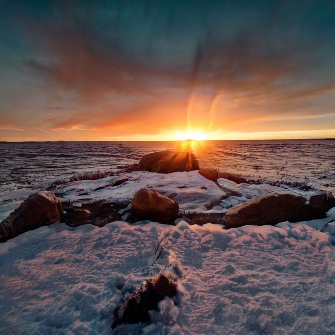 A breathtaking sunset casts golden rays over a snow-covered field scattered with rocks, evoking a peaceful end to the week. The serene, warming horizon pairs perfectly with the uplifting "GM" energy and weekend vibes of the post.