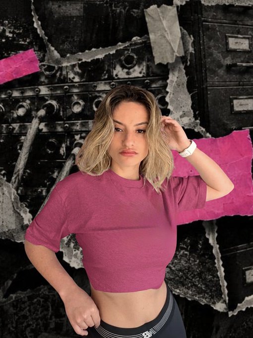 The same image as above - a young woman with blonde hair wearing a cropped magenta/pink t-shirt posing against a black and pink torn paper collage background.