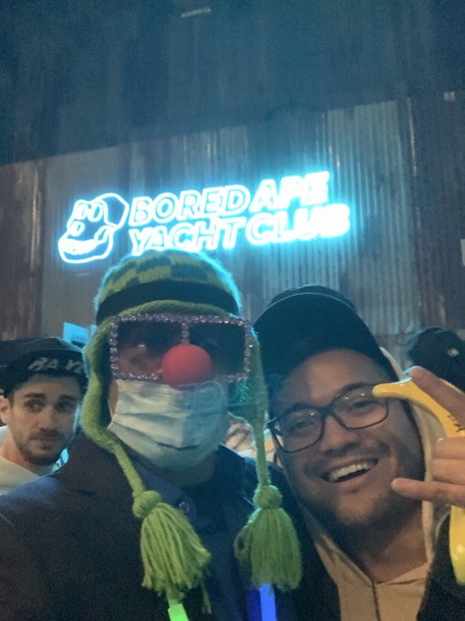 Two people posing in front of a neon "BAYC BORED APE YACHT CLUB" sign; one wears a clown nose, a face mask, and a green knit hat, while the other gives a thumbs-up holding a banana.