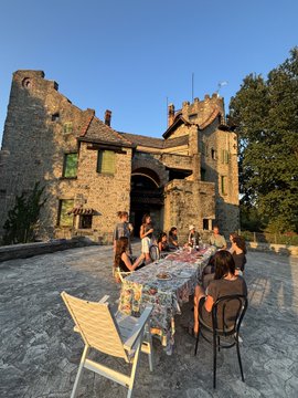 A group of people gathered around a long dining table set up outside a stone castle or historic building at dusk.