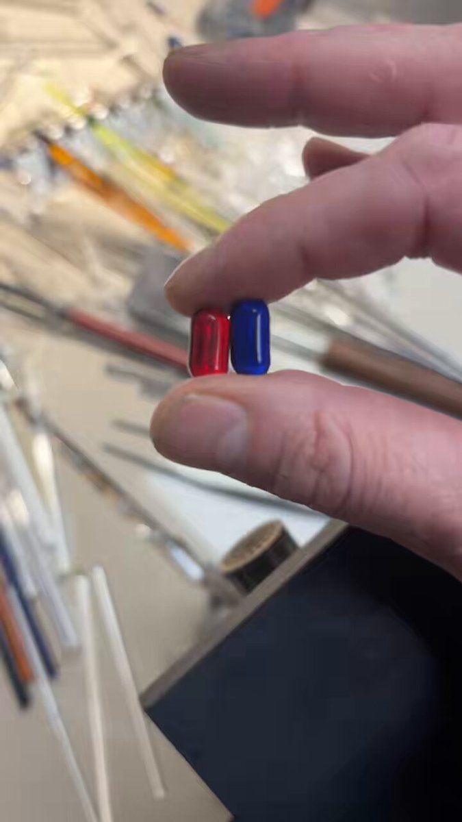 urn of glass rods, a pair of red and blue pills, and a hand holding them up against a gray background.