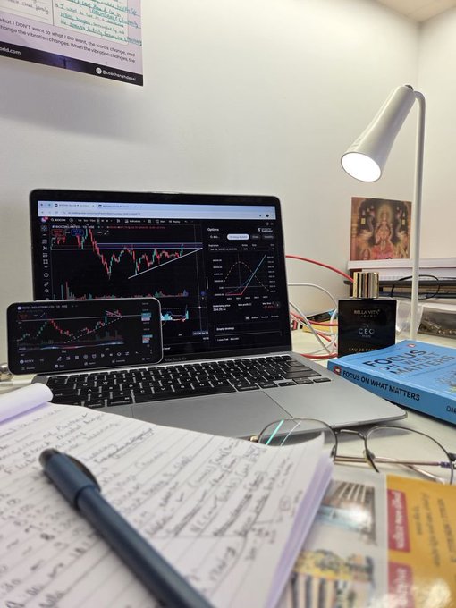 A dedicated trader's workspace features a laptop and smartphone displaying candlestick charts, alongside handwritten notes, a pen, reading glasses, and a "Focus on What Matters" book, reflecting the post's message about embracing risk and persisting through market challenges.