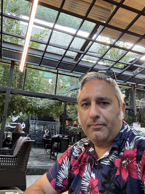 A selfie of a man wearing a dark blue floral Hawaiian shirt and sunglasses perched on his head, sitting at an outdoor cafe with a glass roof.