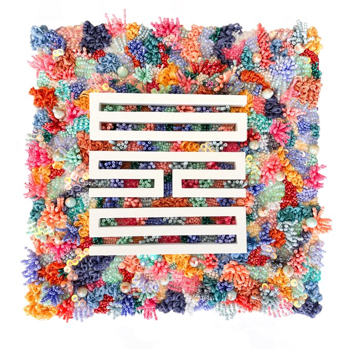 A square mixed-media artwork features a maze-like white maze pattern in the center, surrounded by a dense, textured border made of colorful fabric flowers, beads, and yarn in shades of pink, blue, orange, and green.