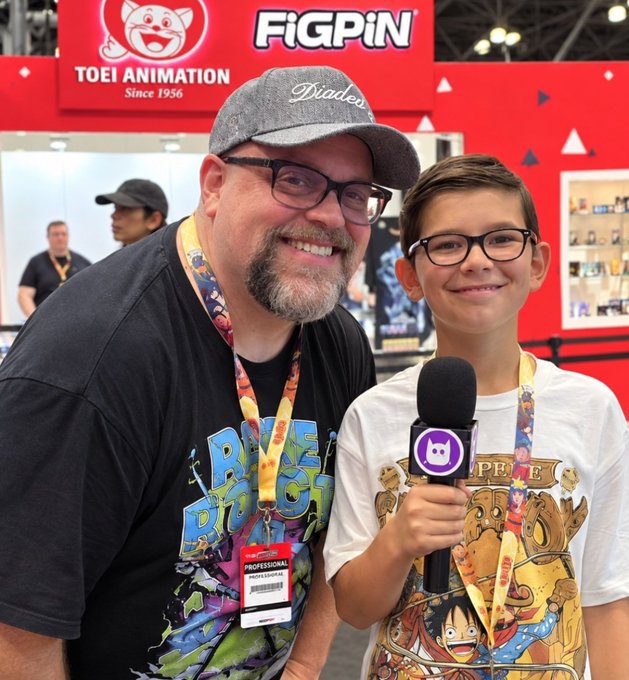 A man wearing a "Dialed" cap and a Black Dragon t-shirt poses with a young boy holding a microphone at a Toei Animation and FiGPiN convention booth.