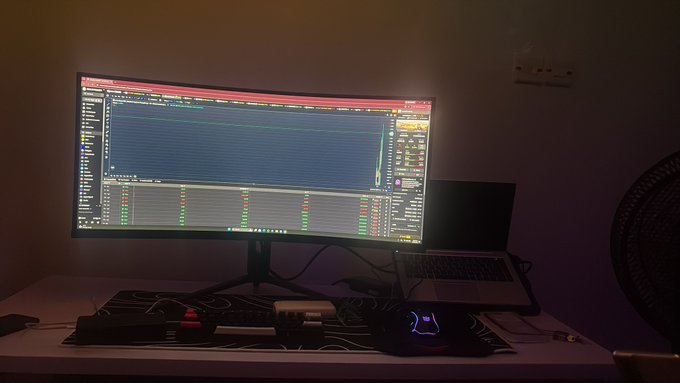 A dimly lit desk setup featuring a curved monitor displaying what appears to be a trading or data analysis software, accompanied by a laptop, mouse, and various accessories.