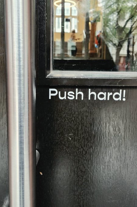 A black door with a glass panel shows the text 'Push hard!' on its lower portion, with reflections of people and trees visible in the window.