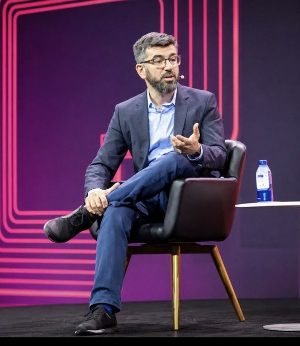 A man with glasses and a beard in a dark blazer and blue shirt sitting in a chair on stage during what appears to be a conference or panel discussion, with a pink neon backdrop.