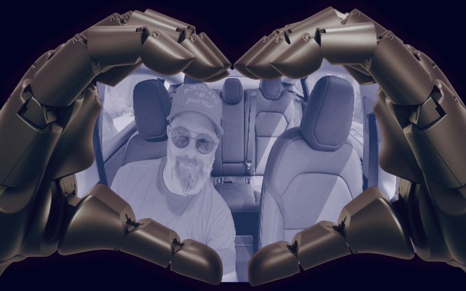 A man with a beard and sunglasses sitting in a car, framed by two large metallic mechanical crab claws forming a heart shape.