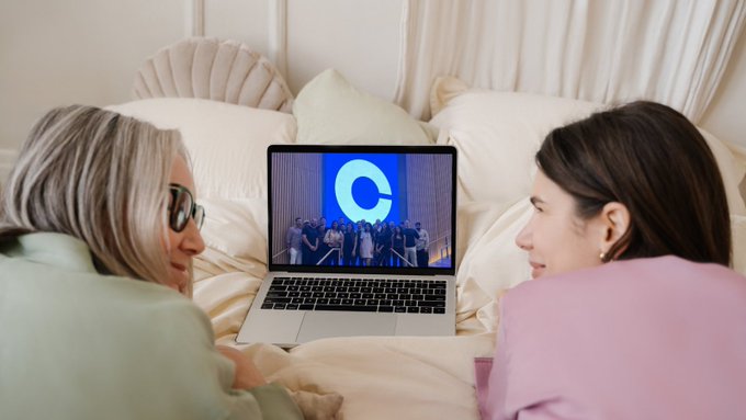 Two women lie on a bed with cream-colored bedding, watching a video on a laptop showing a group of people holding up a large blue 'C' logo.