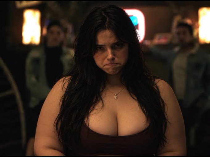 A woman with long dark hair and a maroon top makes a displeased pout in a dimly lit bar setting.