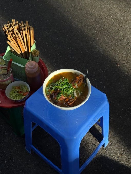 A bowl of noodle soup with greens and meat served on a small blue plastic stool on a street, accompanied by a condiment tray with chopsticks and sauces.