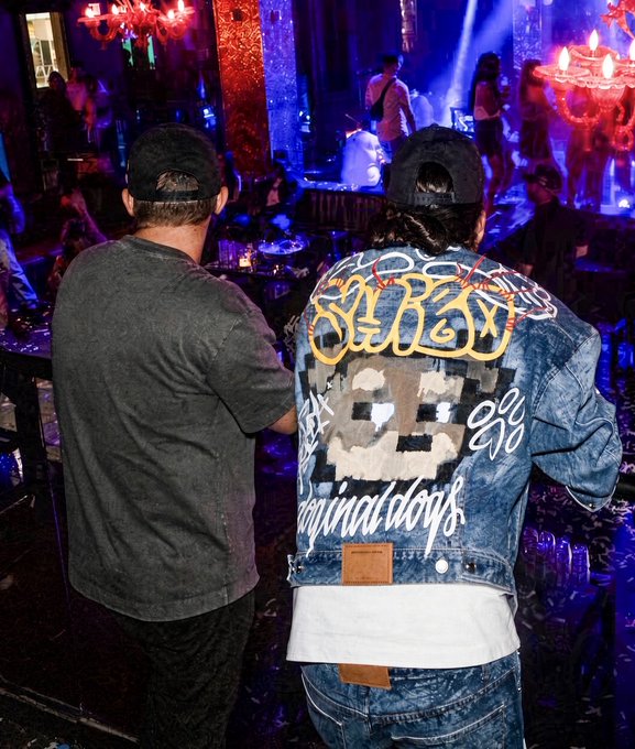 Two people stand in a dimly lit club setting with chandeliers, one in a gray shirt and the other wearing a denim jacket with elaborate graffiti-style artwork on the back.