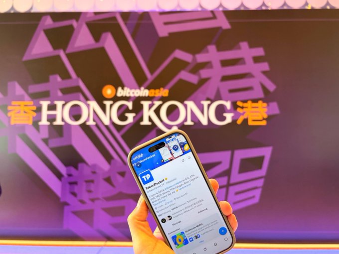 A hand holds a smartphone displaying a social media feed in front of a purple backdrop featuring the 'HONG KONG' text and 'Bitcoin Asia' branding.