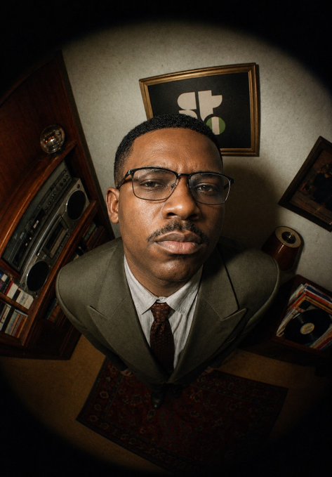 A fisheye-lens selfie of a man wearing glasses and a green suit, standing in a room featuring a vintage boombox, framed artwork, and a Persian rug.
