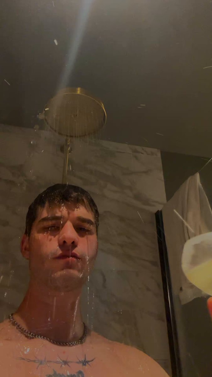 The image shows a man standing under a shower head, with his face partially visible above the water spray. The background of the photo features a bathroom setting, including a marble wall and a towel hanging nearby. There are also two cups present in the scene - one on the floor next to the man's feet and another further away from him.