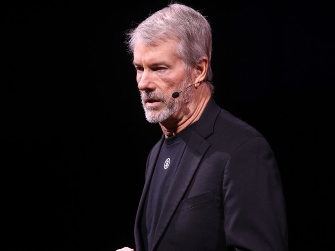 Michael Saylor, wearing a black blazer and headset microphone, speaks at a podium during a presentation or interview. The image relates to the post as it shows Saylor, co-founder of Strategy, who endorsed the CLARITY Act to provide regulatory clarity for digital assets.