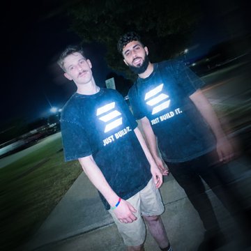 Two young men wearing matching dark t-shirts with 'JUST BUILD IT.' printed on them, posing together outdoors at night.