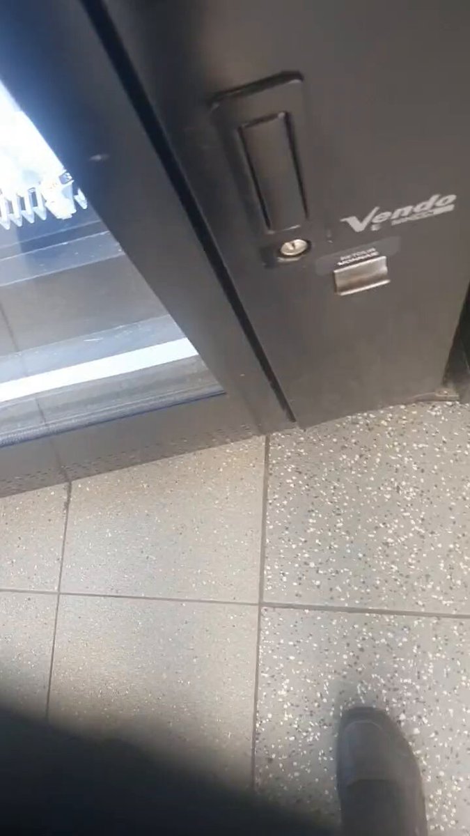 The image shows a person's feet with black shoes, standing next to a door that has a Vendo logo on it. The floor appears to be tiled, and there are two people visible in the scene - one near the left side of the frame and another closer to the right edge.