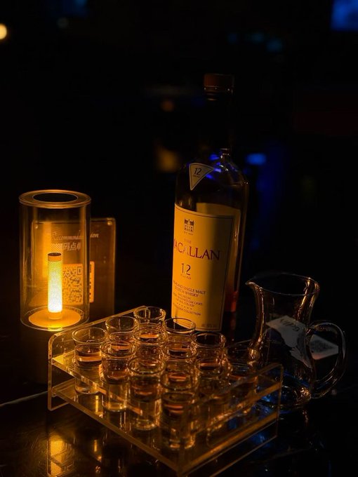 A warmly lit bar scene featuring a bottle of **Macallan 12** Single Malt Scotch Whisky surrounded by neatly arranged tasting glasses and a decorative candle lamp. The image evokes premium craftsmanship and tradition, contrasting with the post's discussion of stability and infrastructure upgrades in TermPrime's Canton rollout via CIP-0119 and CIP-0112.