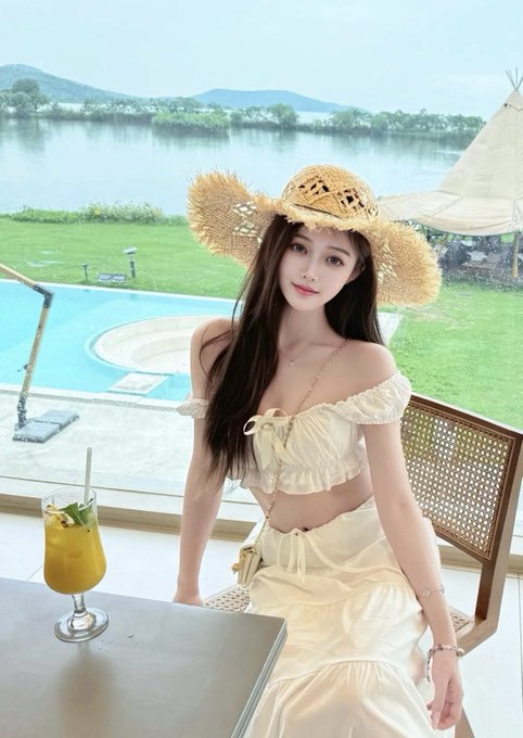 A young woman in a straw sunhat and white outfit sitting at a table with a tropical drink, overlooking a pool and lake scenery.