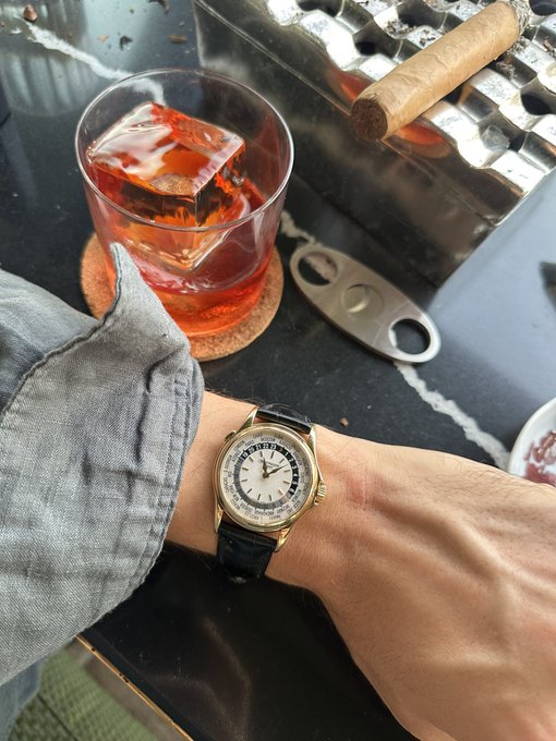 A man's wrist displays a yellow gold Patek Philippe World Time watch (reference 5110J) with a guilloche dial and black leather strap, photographed against a black marble surface alongside a glass of reddish-brown cocktail on a coaster, a metal cigar cutter, and a cigar resting on a foil-wrapped tray. The watch features the signature world time city dial encircling a guilloche center, matching the post's description of this newly acquired dressier timepiece.