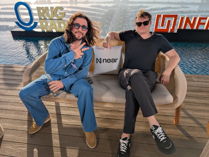 Two men sit on a beige couch outdoors by the water, with 'RUG RADIO' and 'INF' logos visible in the background and a pillow labeled 'near' on the couch.