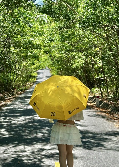 A person in a white dress walks down a sunlit tree-lined path holding a bright yellow umbrella branded with "COIN98" logos. The image visually matches the post's announcement about the exclusive Coin98 umbrella merch being available at the Coin98 booth at Conviction VN 2026.