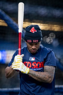 A baseball player in a Boston Red Sox beanie and navy shirt holds a bat over his shoulder, wearing a yellow and white batting glove with visible arm tattoos.