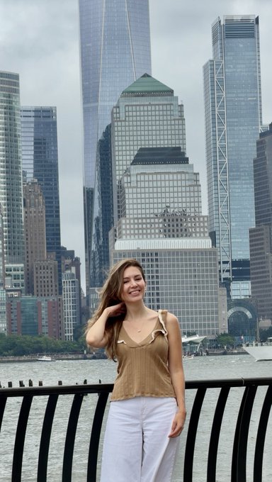 A young woman with long brown hair stands smiling on a waterfront promenade, wearing a beige ruffled top and white pants, with a backdrop of tall skyscrapers including One World Trade Center.
