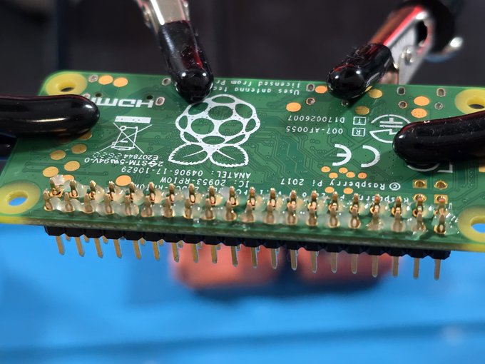 A Raspberry Pi Zero board is held by a pair of helping hands, its golden GPIO pins prominently displayed with markings including "Raspberry Pi 2017" and "IC: 20953-DPIOW" visible on the underside, fitting the "creamy pins for added entropy" caption as the exposed headers appear ready to add randomness to any project.
