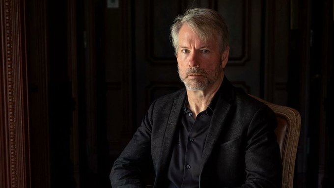A professional portrait of a gray-haired man with a beard wearing a dark blazer, seated in a dimly lit wood-paneled room.