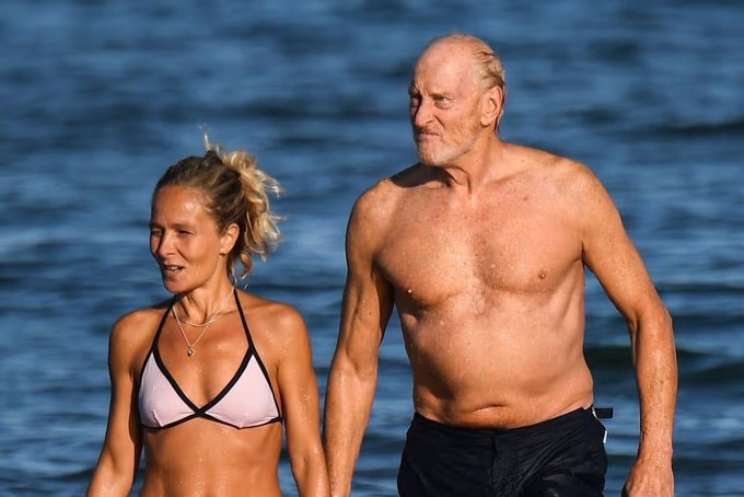 An older shirtless man with grey hair walking on a beach alongside a woman in a pink and black bikini top, both appearing wet from the water.