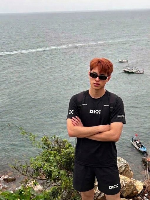 A young man with reddish-brown hair and sunglasses stands confidently with crossed arms at a coastal cliff, wearing a black "OKX" branded t-shirt and matching shorts with a backpack, overlooking the sea with small boats visible in the water below—showcasing the OKX merchandise set he's reviewing after returning home.