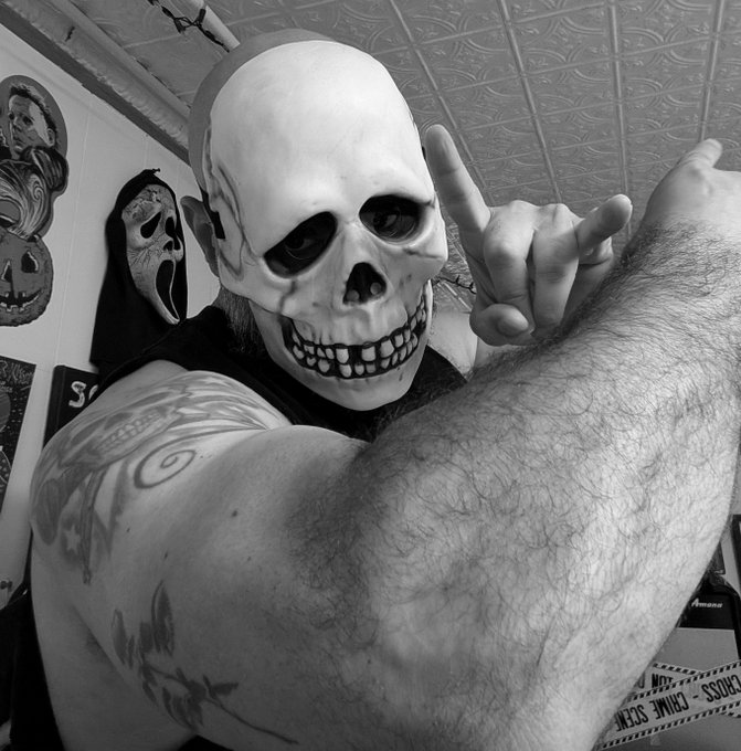 A black and white photo shows a person wearing a skull mask, flashing a hand gesture, with visible tattoos on their arm and horror masks in the background.
