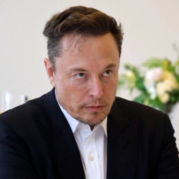 The image features an adult male wearing a suit, with his head tilted slightly to one side as he looks off into the distance or at something out of frame. The background includes a white wall and some flowers, adding a touch of color and life to the scene. There is also text in the image that reads "Elon Musk: Bitcoin is a good thing." This suggests that the man might be discussing or contemplating the impact of Bitcoin on society or technology.