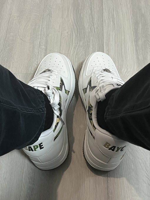 A first-person view of someone wearing white, black, and camo BAPE sneakers standing on a wooden floor.