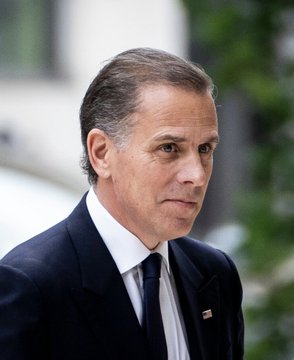A middle-aged man in a dark suit and blue tie, wearing an American flag lapel pin, stands outdoors with a serious expression, appearing to be Hunter Biden. The image accompanies a post noting that Hunter Biden's cryptocurrency "$LAPTOP" has plummeted 99% from its launch value, with its market cap falling under $100 million.
