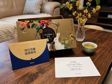 A mooncake gift set from Huobi (火币网) sits on a coffee table alongside a greeting card reading "且行万里 踏阅有光" and a small teacup of tea, with the user having carefully kept last year's "May" card displayed alongside this year's. The arrangement reflects the user's sentiment of treasuring these annual greeting cards from Huobi, showing both a current mooncake box and a previously saved card in a warm, contemplative scene.